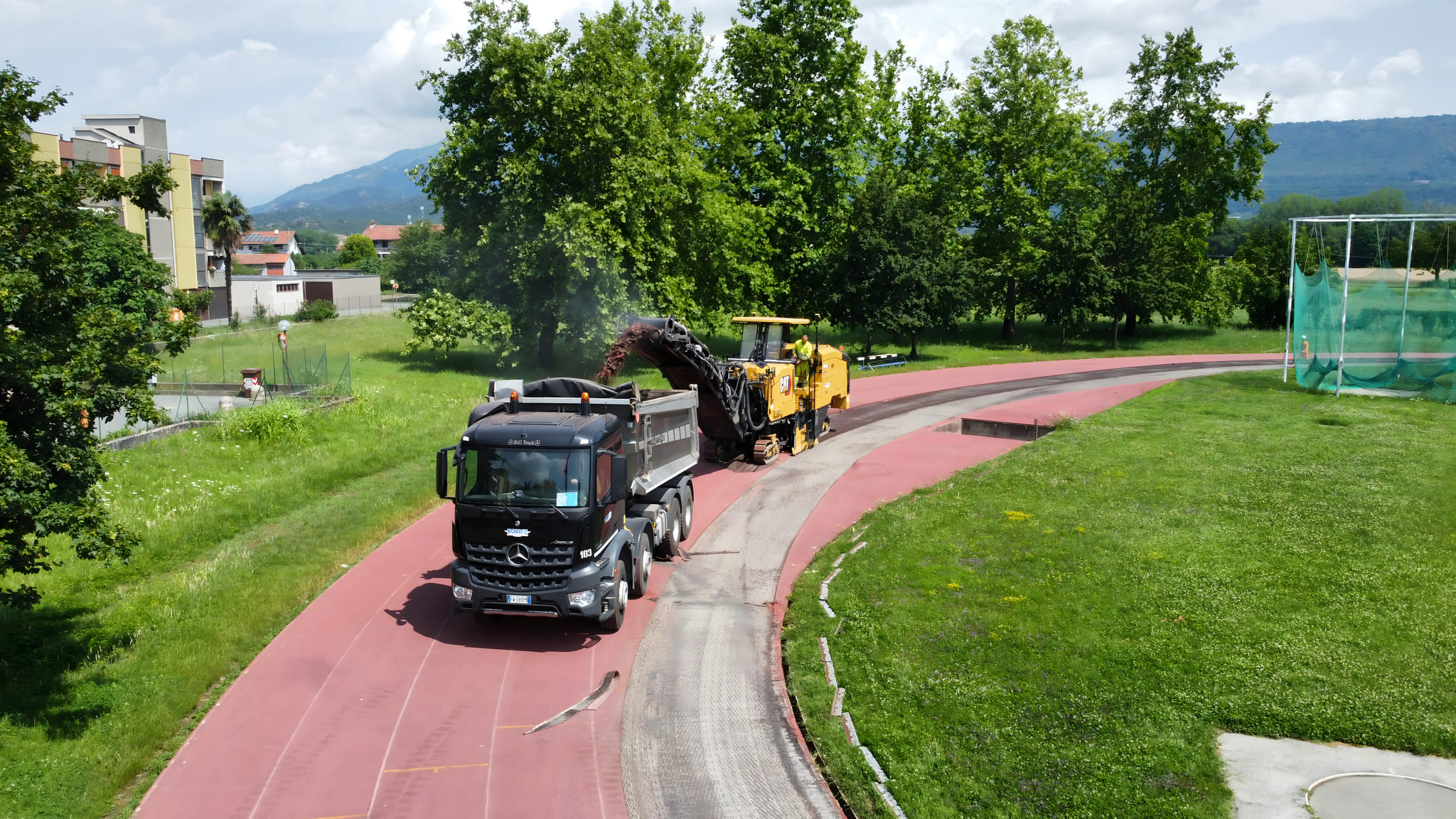 Fresatrice stradale CAT in azione sulla curva di una pista di atletica in tartan rosso, con camion ribaltabile Mercedes-Benz Arocs nero in fase di raccolta del fresato durante lavori di rifacimento del manto sportivo