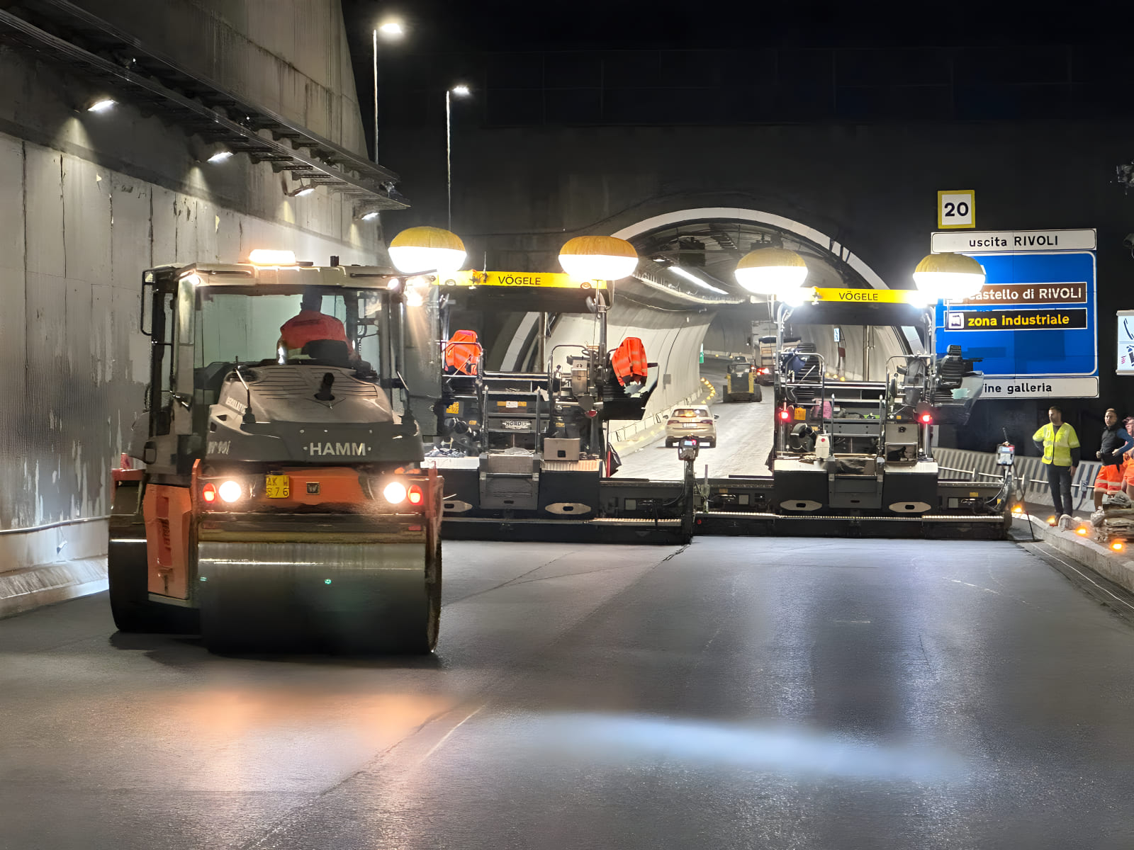 Rullo compressore Hamm e due finitrici stradali Vögele in tandem durante lavori notturni di rifacimento del manto stradale all'imbocco della galleria autostradale di Rivoli, con segnaletica autostradale visibile sullo sfondo
