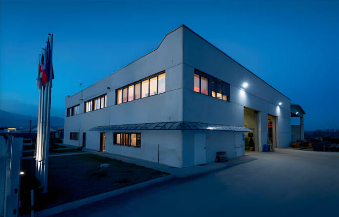 Vista esterna notturna della sede operativa di Nordex SpA, edificio industriale a due piani con bandiere istituzionali e illuminazione esterna