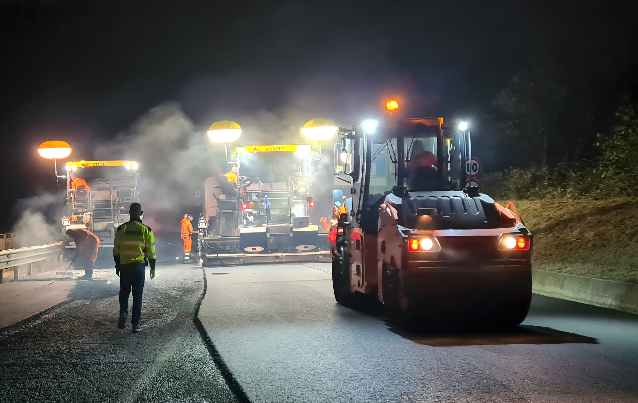 Cantiere notturno di rifacimento del manto stradale con due finitrici Vögele in tandem e rullo compressore in primo piano, operai in tuta ad alta visibilità tra vapore dell'asfalto caldo