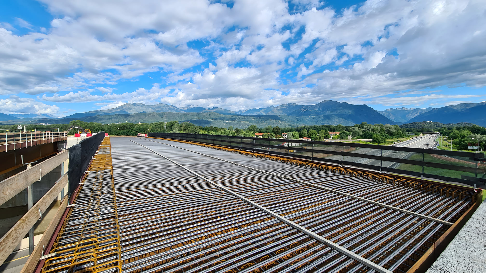 Vista prospettica dell'armatura in acciaio con rete di tondini posata sull'impalcato di un viadotto autostradale in costruzione, con paesaggio alpino e autostrada visibili sullo sfondo